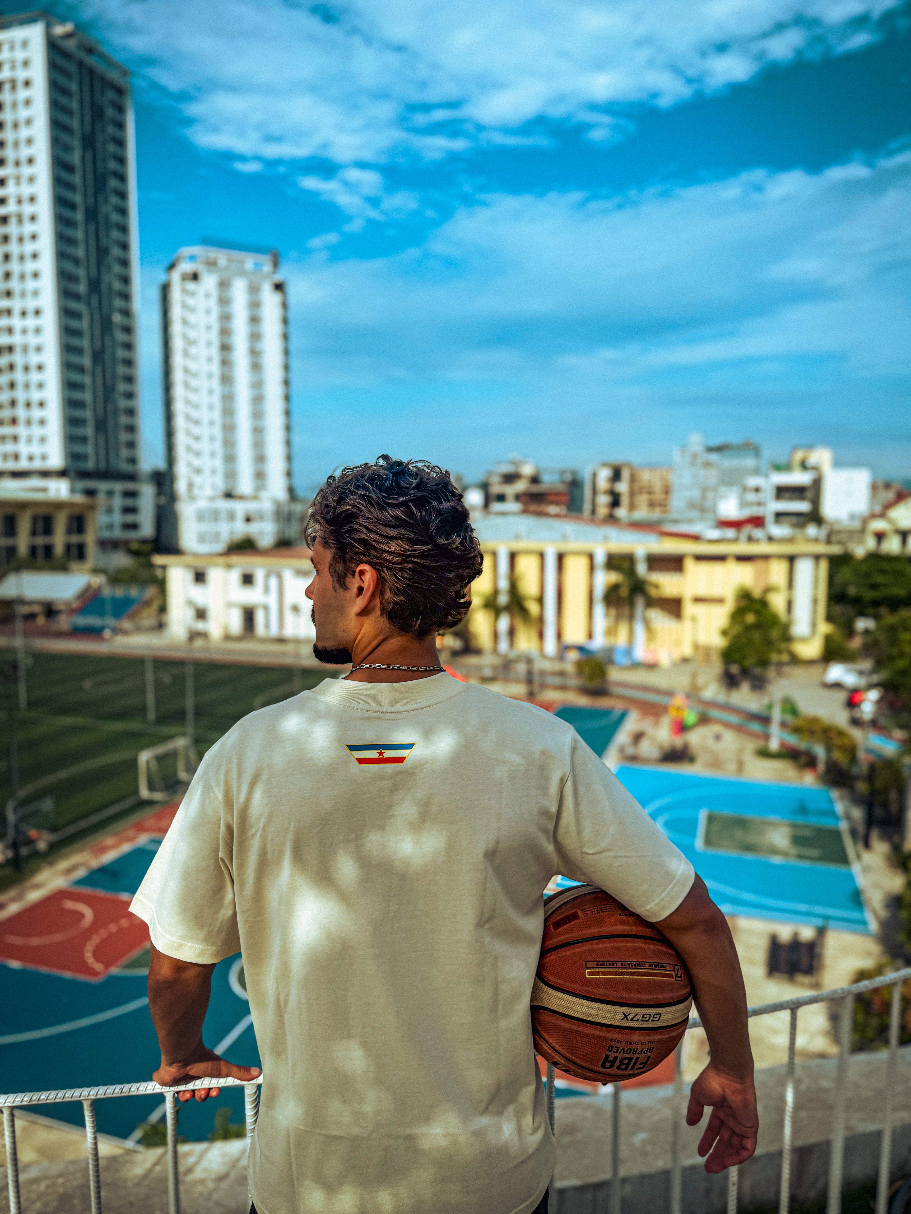 Man showing the back of a Kin and Crest Ivory Team Yugoslavia Basketball Tee holding a basketball on a rooftop overlooking basketball courts with buildings and a clear sky.