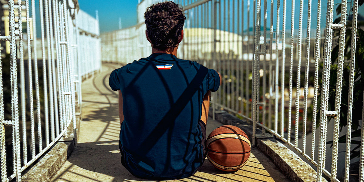 Man sitting on a bridge with a basketball, showing the back of a Navy Kin and Crest Team Yugoslavia Basketball Tee.