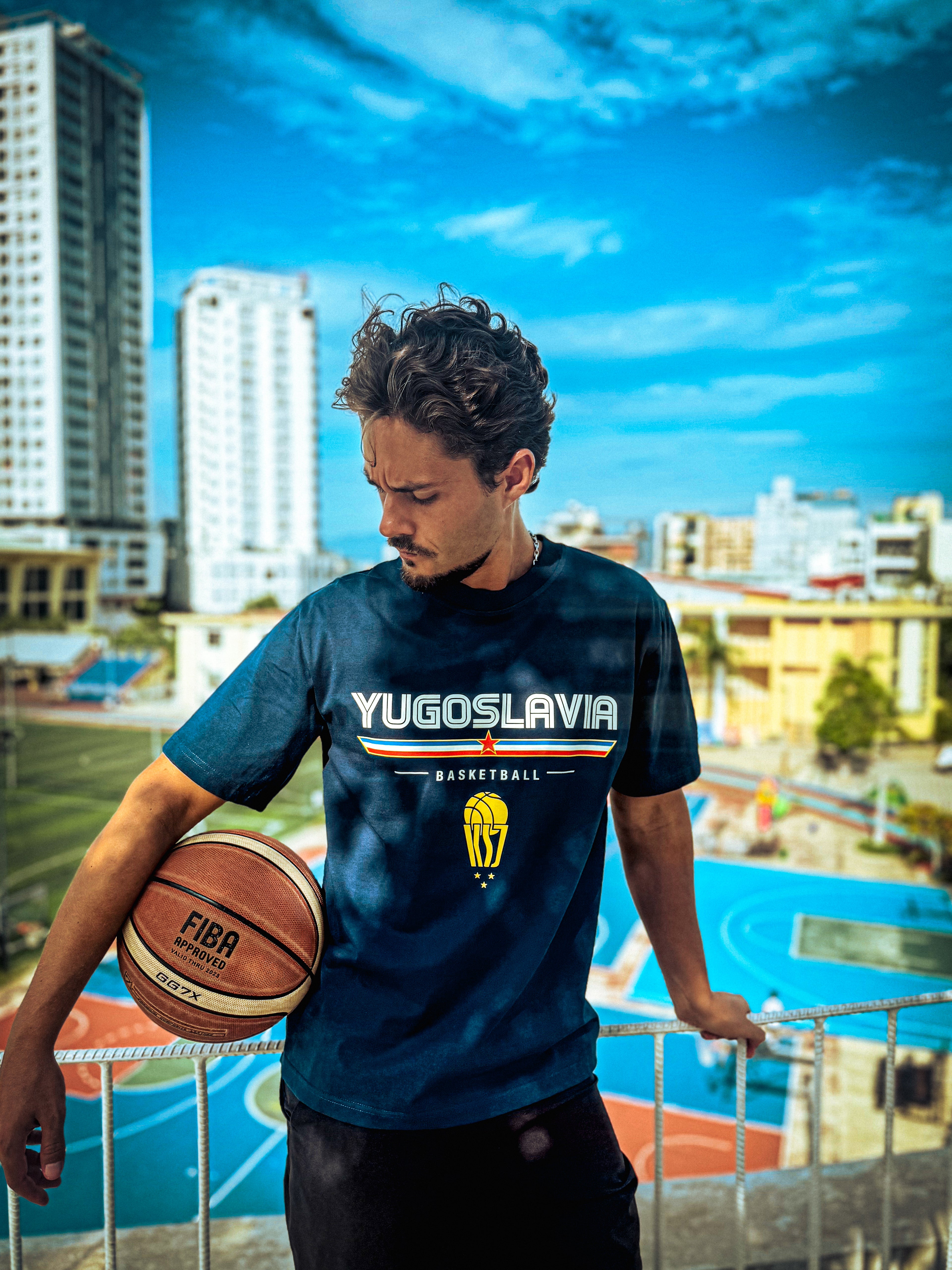 Man holding a basketball wearing a Navy Kin and Crest Team Yugoslavia Basketball Tee with basketball courts in the background.